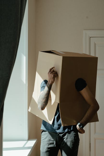 A person with tattooed arms wearing a black watch and black gloves is carrying a large cardboard box during a home relocation process inside a room with beige walls and a white window frame. The indiv