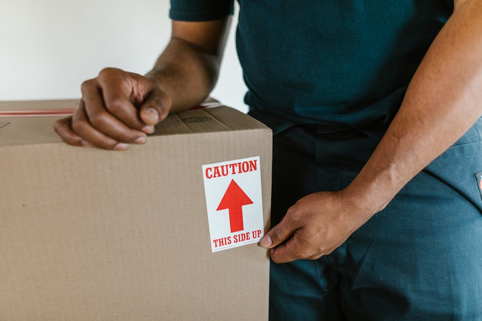 A person with tattooed arms wearing a black watch and black gloves is carrying a large cardboard box during a home relocation process inside a room with beige walls and a white window frame. The indiv