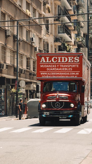 A red Mercedes-Benz moving truck with a large sign on top advertising Alcides, a company specializing in furniture transport and cargo services, parked on a city street in front of multi-storey residential buildings with balconies and potted plants. The front of the truck faces the camera, with the Mercedes logo visible on the grille. Nearby, a person is standing on the pavement near a grey waste bin, while another individual appears to be walking past the truck. The street is shaded by the buildings, and the scene suggests a home relocation or moving process taking place in an urban environment, with the truck poised to load or unload furniture and boxes for a house removal or moving service, as part of a professional packing and moving operation facilitated by Removal Companies Earls Court.