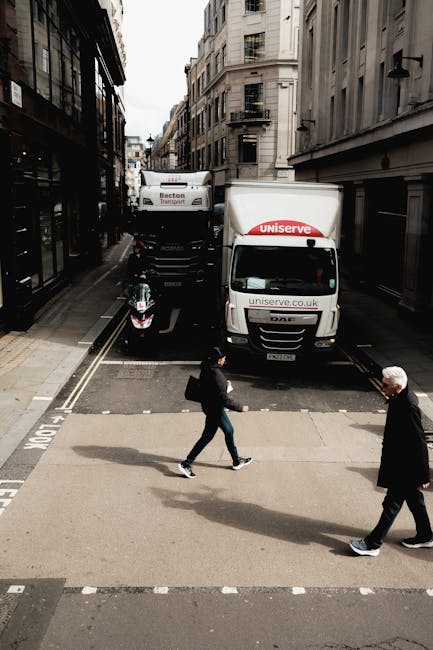 A street scene on a narrow urban road with tall, multi-storey buildings on either side, featuring two vans parked parallel to the curb with visible branding, one marked 'Uniserve' and the other 'Becton Transport.' A black and pink motorcycle is parked next to the vans, close to a roadside loading area. In the foreground, two pedestrians are crossing the street on a zebra crossing; one is a woman carrying a backpack and wearing casual clothing, while the other is an older man dressed in dark clothing. The buildings have fire escapes and balconies, with a cloudy sky overhead. The scene depicts a typical city environment during a home relocation process, with the presence of vehicles and pedestrians engaged in some form of moving or transport activity, relevant to house removals and logistics handled by Removal Companies Earls Court.