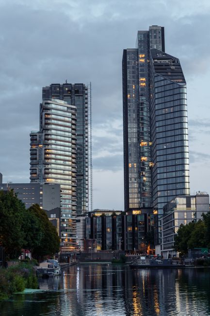 The image displays a cityscape featuring several modern high-rise buildings with glass facades reflecting the evening sky, situated along a waterway. In the foreground, there are leafy green trees and a small dock with boats moored beside it. Inside a residential property, visible through an open doorway or window, there are cardboard boxes, some wrapped in plastic or packing paper, stacked on the floor or being carried by individuals during a home relocation. Furniture items such as a small table or chairs are partially visible, along with moving blankets and hand trucks used for furniture transport. The scene captures the process of loading or unloading household items as part of a professional removals service, possibly provided by Removal Companies Earls Court, supporting efficient packing and moving logistics for London flats like those referenced in the page about Lillie Square apartment removals.