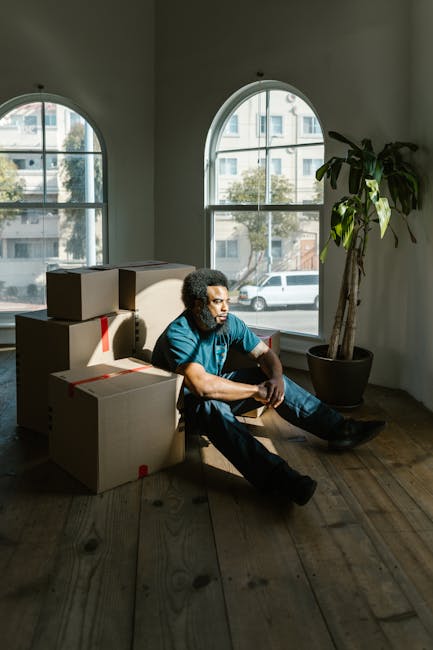 A man with curly dark hair and medium skin tone, wearing a blue T-shirt and dark trousers, is seated on a hardwood floor inside a room, leaning against a beige sofa surrounded by cardboard moving boxes sealed with red tape. The boxes vary in size and are stacked in a semi-organised manner nearby. The room has two large, arched windows with black frames, allowing natural light to illuminate the space and revealing an urban street scene with parked cars and buildings outside. To the right, there is a tall potted plant with broad green leaves, contributing to the room's interior decor. This scene depicts the process of home relocation, with furniture and packing materials indicating ongoing packing or unpacking, in a setting consistent with house removal services provided by [COMPANY_NAME].