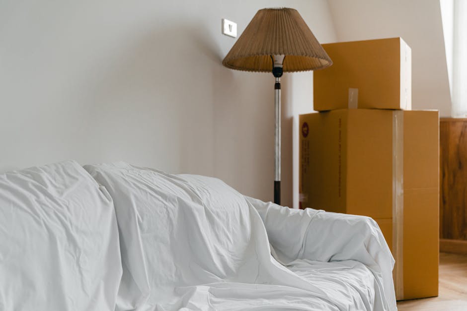 Inside a room prepared for house removal, there are three cardboard moving boxes stacked next to a wooden cabinet near a window. A floor-standing lamp with a brown shade is positioned behind the boxes. To the left, a white sofa with a slightly wrinkled cover is visible, occupying most of the foreground. The door frame can be seen behind the boxes, indicating the area is part of a home interior. The natural light coming through the window illuminates the scene, highlighting the packing and furniture ready for transport by a professional removals service such as Removal Companies Earls Court. This image illustrates typical home relocation preparations involving packing, furniture transport, and loading processes.