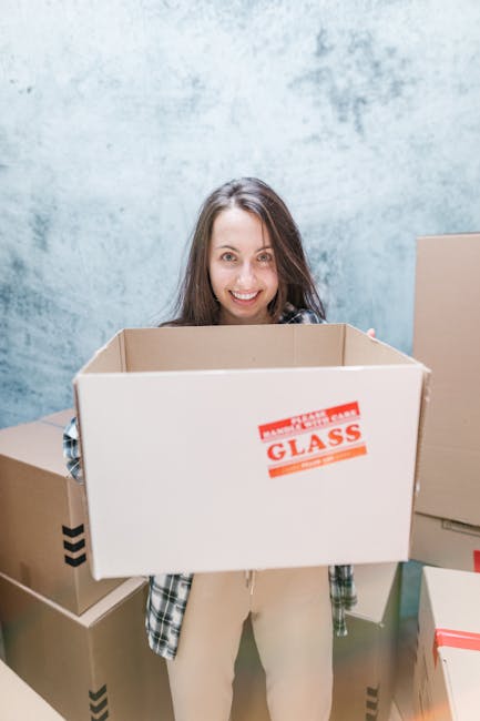 A young woman with long dark hair, smiling, standing inside a property surrounded by several cardboard moving boxes. She is holding an empty, open cardboard box labelled 'Fragile - Handle with Care - Glass' in red text, indicating careful packing for home relocation. The background features a light blue textured wall, and additional boxes of various sizes, some sealed with packing tape and others open, are positioned on either side of her, suggesting a packing or moving process. The scene is lit with natural or soft indoor lighting, reflecting a professional and organized approach to furniture transport and packing for a house move, consistent with services offered by Removal Companies Earls Court.